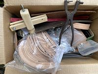 Top view of a box showing a wooden clamp tool, metal hole punch plier, a metal stitching groover and a plastic bag of rolled leather strips along with other scattered pieces and rivets.
