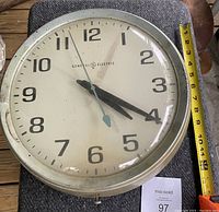 General Electric 13 inch diameter clock face with visible wear, showing brand name, black Arabic numerals, and hour/minute hands into the 4 o'clock position with a measuring tape confirming size.