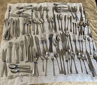 Full wide shot of laid out flatware on white cloth showing many pieces of stainless steel knives, forks, spoons, and other utensils arranged by type and size.