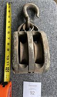 Top view of the vintage pulley block showing two large sheaves and metal housing next to tape measure indicating size approximately 9 inches vertical length including hook.