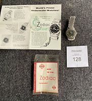 Front view of two Zodiac wristwatches on stainless steel beads-of-rice bracelets alongside original Sea Wolf advertisement flyer and sealed "How to Use Zodiac Rotographic" manual, with lot tag visible.