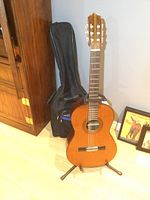 Full view of the Zen-On acoustic classical guitar on its stand with the black soft-sided carrying case behind, showing the wooden body and neck, placed on light wood floor near a wooden cabinet.
