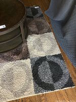 Photo showing the synthetic fibre area rug with a geometric pattern, brown round table partially on it and a curtain beside it.