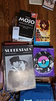 Top view of five assorted books including 'The Mojo Collection', 'No One Here Gets Out Alive' biography of Jim Morrison, 'Superstars' on American rock stars, and a vinyl record in sleeve.