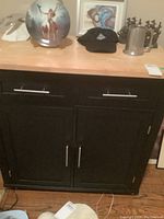 Front view of the black cabinet showing two doors and two drawers with silver handles, butcher block top with several items on it.