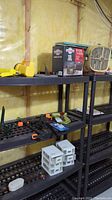 Wide shot showing black shelving unit with Motomaster battery charger box, tabletop fan, clamps, caulking tool, and patio chair cover on the shelves against an unfinished basement wall.