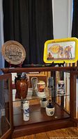 Wide shot showing the Coca-Cola tray, ceramic steins, glass plate, and Delft bottle displayed inside a glass cabinet