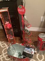 Photo showing metal bottle holder with four clear glass Coca-Cola bottles, metal waste bin, boxed Castline truck and Coca-Cola can light, playing cards, flashlight, and red vertical tower.