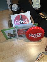 Set of Coca-Cola and related advertising items including metal bottle cap, wood crate, and paper ads on a table
