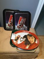 Four black rectangular trays stacked upright featuring a red and white Coke can on ice and one oval tray below them.
