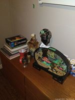 Photo shows household items on top of a wood cabinet, including Jim Beam eagle decanter, skull light, oval oriental art display, duff bottle opener, stacked books, and visible ends of the Beatles light and Terminator figure box.