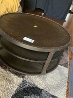 Front side angle view of round drum style coffee table showing metal top and two wooden shelves with rivet details on metal frame