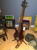 Full view of Cort electric bass guitar standing on a black tripod stand with brown leather strap attached, surrounded by decorative items and posters.