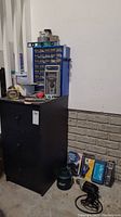 Wide view of the black storage box with small parts organizer, various measuring tapes, screwdriver sets and other small hardware items on a concrete floor against a white and brick wall.