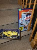 Photo of a yellow die cast Pepsi-Cola truck bank in original box placed on stairs next to a vintage style Pepsi-Cola metal sign with a fountain advertisement and glass promo, sign size about 12x17 inches.