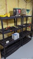 Two black resin shelving units, matching design, shown with various items placed on the shelves for size reference. Shelves have four tiers each, with slatted surfaces.