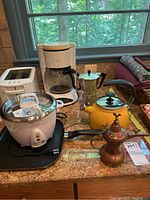 Photo of all items including Kenmore toaster, Mr. Coffee maker, Aroma rice cooker, yellow tea kettle, metal coffee maker, and copper incense burner on countertop near window.