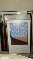 Several framed posters and art pieces stacked upright against a wall, front visible poster is the Santa Fe Chamber Music Festival.