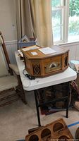 Photo of white folding catering table with items including a wooden decorative radio on top, and some papers and upholstery chair beside it, showcasing the table's dimensions and condition.