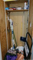Wooden cabinet containing mop, broom, bucket, bottles of cleaning liquids and cans on wooden shelves, and laundry basket below filled with tote bags