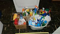 Overview of the cleaning supplies lot showing various spray bottles, detergent containers, and tools in cardboard boxes and plastic bins.