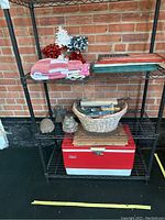 Overall view of Coleman cooler, gardening tools in basket, towels, decorative stars, and hanging signs on metal shelving against brick wall.
