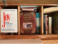 Top view of the cookbooks including The Joy of Cooking, The Original White House Cookbook, and other books in a wooden shelf compartment