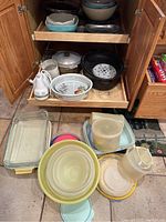 Photo of various cookware items inside kitchen cabinet and on floor including bowls, roasting pans, casserole dishes, and Tupperware containers.
