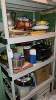 Wide view of multi-tier shelving unit showing cookware, bakeware including cake pans, a blender, thermoses, and food storage items stacked on multiple shelves in basement storage.
