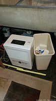 Comfort-Aire dehumidifier and beige plastic trash can beside each other on the floor.