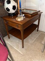 Front and side view of the wooden end table showing the single drawer with metal ring pulls, lower shelf, and tapered legs, with various decorative items placed on top of it.