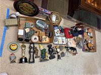 Top-down view showing assorted figurines including golf gnome, trophy, bird figures, metal belt buckle, and other collectibles arranged on floor with some items in box.
