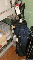 Photo of blue golf bag with clubs standing upright on floor by shelf, tags attached to bag and clubs, clubs include some with headcovers.