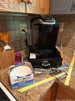 Photo of black Keurig coffee maker on kitchen counter with instruction booklet and wooden holder on side, including box of Mr. Coffee coffee filters, vintage hand mixer beater attachments, yellow kitchen tongs and baster visible.