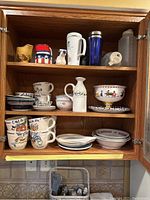 Kitchen cabinet with assorted mugs, a white ceramic pitcher, bowls, plates, and plastic water bottles.