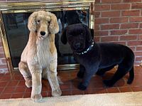 Two life-size plush dogs, tan and black, seated on floor in front of fireplace