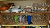 Shelf displaying clear glass stemware (wine glasses and flutes) stacked paper plates, napkins, plastic cups, napkin rings and woven baskets.