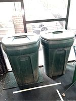 Pair of Rubbermaid trash cans with lids, wheels, and dirty exterior, shown indoors by glass door.