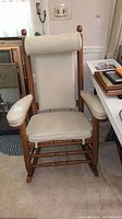 Full view of vintage upholstered rocking chair with beige cushions on seat, armrests, backrest and cylindrical pillow headrest. Wooden frame with natural finish and round finial posts.