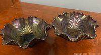 Two identical iridescent carnival glass candy dishes side by side on a wooden surface. Shows the ruffled edges, iridescent finish, and embossed center pattern.