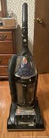 Front view of black upright Hoover vacuum cleaner on wooden floor near furniture and door.