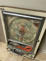 Full view of the Japanese pachinko game machine showing the wooden frame, cracked glass front panel, circular playing field with pins, and front control panel with lever and ball tray.