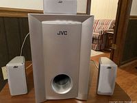 JVC home theater sound system with a large silver subwoofer and three matching smaller speakers placed on a brown floor with sofa in background