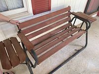 Front and side view of outdoor wooden rocking bench showing horizontal slats, metal frame, armrests with attached side tables, and rocking base.