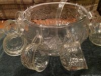 Clear crystal punch bowl with handle, eight matching cups hanging from its rim, a ladle inside the bowl, all placed on a dark surface.