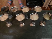 Set of 8 silver goblets arranged on black surface, showing top inside and reflective silver surfaces, with some tarnish visible.