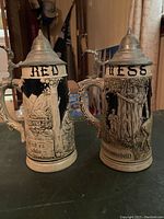 Two German-style ceramic beer steins with detailed relief designs and metal lids on a table.