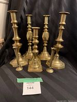 Photo of 8 brass candle holders in various sizes displaying ornate midsections, hexagonal and circular bases, all arranged against a black fabric backdrop.