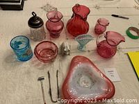 Photo showing cranberry glass pitchers, bowls, creamers, a blue glass bowl, clear jar with metal lid, small silver utensils, and other vintage dishes.