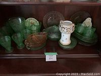 Shelf with various green and pink depression glass pieces, a white ceramic decorative item with gold accents, and a small ceramic dog figurine.